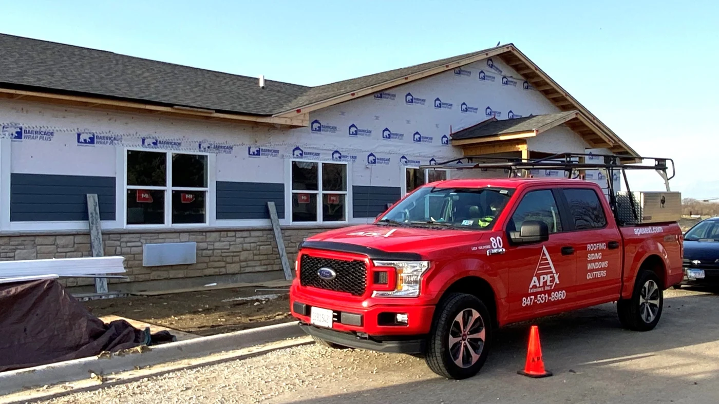 A red Apex Exteriors work truck sits outside what looks like a long commercial building, where Apex teams are working on installing stone veneer skirting and new siding systems.