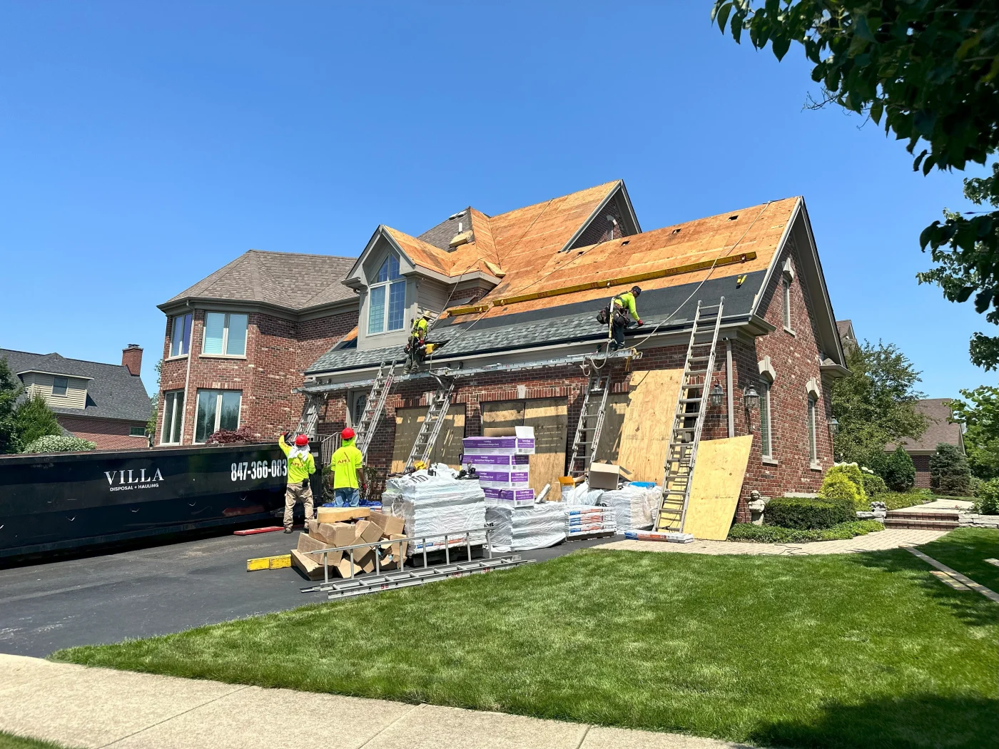 Apex Exteriors Sends Skilled, Trained In-House Exterior Remodeling Crews A beautiful brick home in the Chicagoland area is getting a new light gray shingle roofing system installed by an Apex Exteriors roofing crew, showing them in the process, working on a section of the roof, with some men up on ladders wearing harness and safety gear, and others in the driveway with a dumpster and materials.