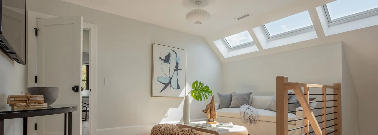 A wide image of a furnished loft with white walls, a painting, couch with pillows, table, and three Velux Skylights flushing the space with natural light.