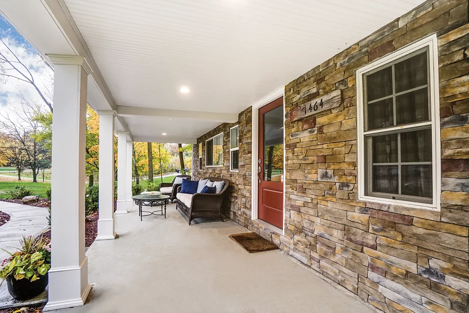 The outside of a wide residential entryway or patio showing a single-entry door and four windows along the length which is covered with rustic engineered stone panels. The walkway is smooth concrete and the ceiling is white with white pillars. There’s some patio furniture including a small table, chair, and couch with pillows.