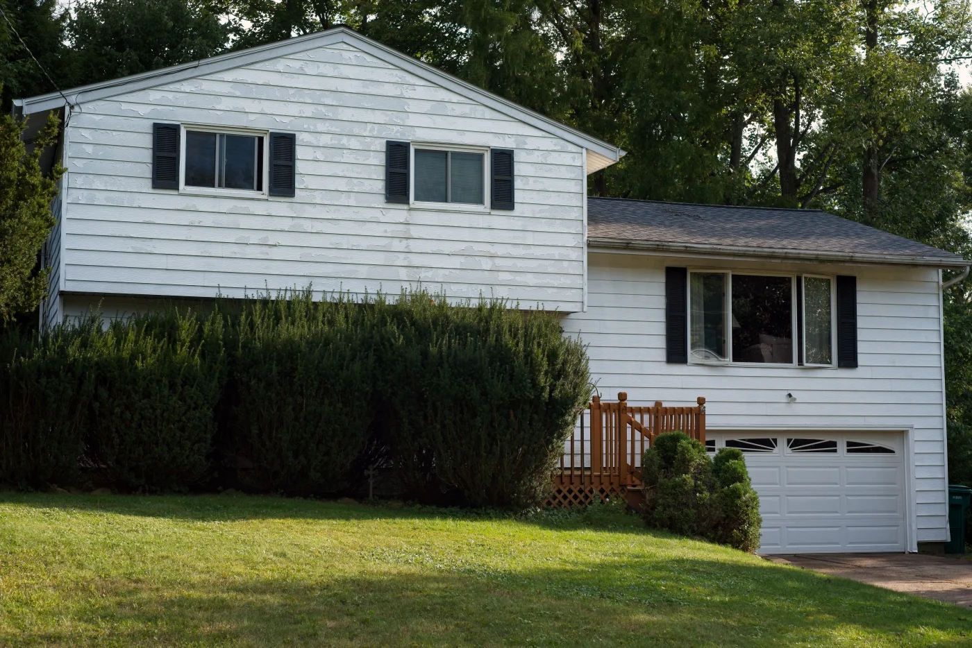 House Peeling Paint A split-level home with a background of trees behind it, with an old roofing system and old siding showing plenty of wear and tear, with repairs likely needed.