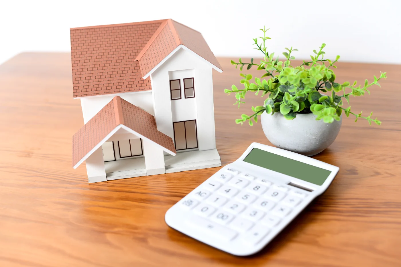 A small miniature home with white walls, and brown shingle roofing on a wood table next to a plant in a small white planter and a calculator, symbolizing an estimate for exterior remodeling work.