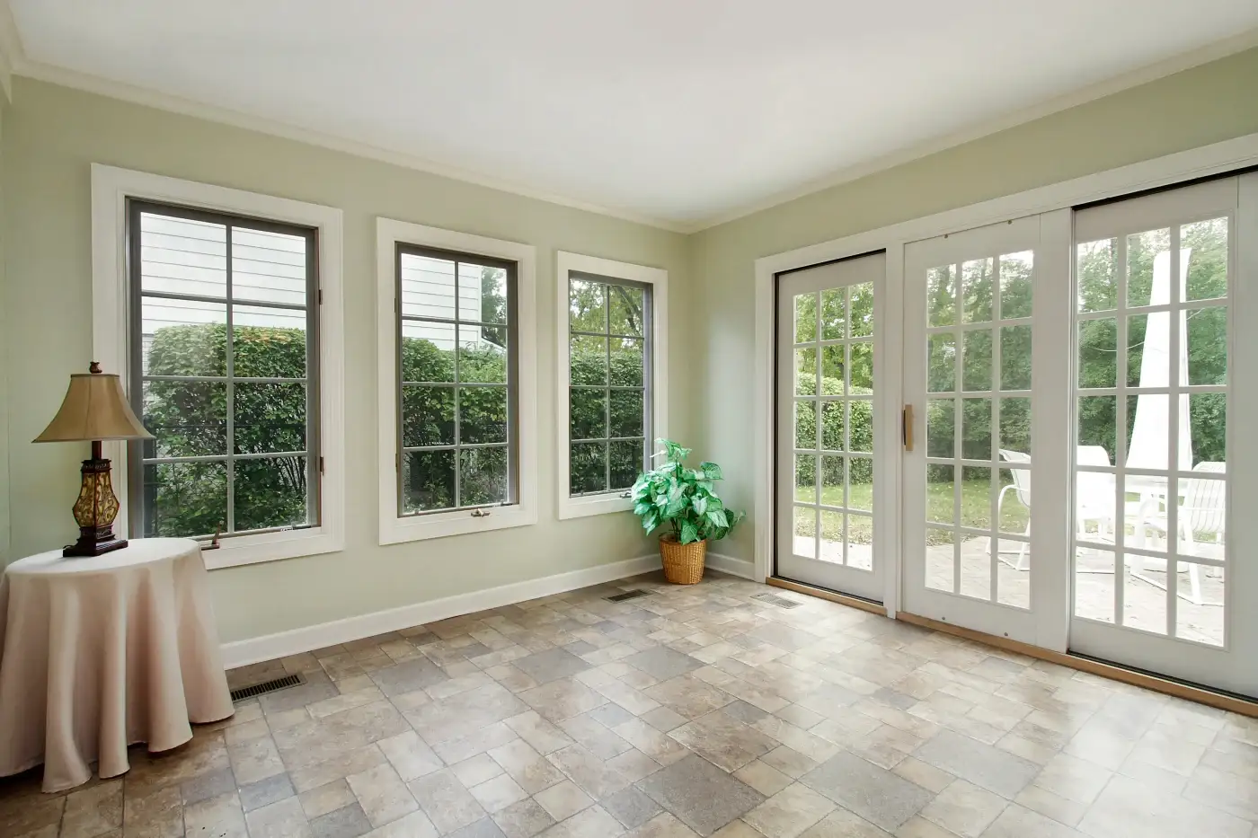 A large open living space in a residential home ready to be moved into, with newly painted beige walls, white trim, white window frames, three picture windows with traditional grids, a single sliding entry door and what looks like a stone tile floor.