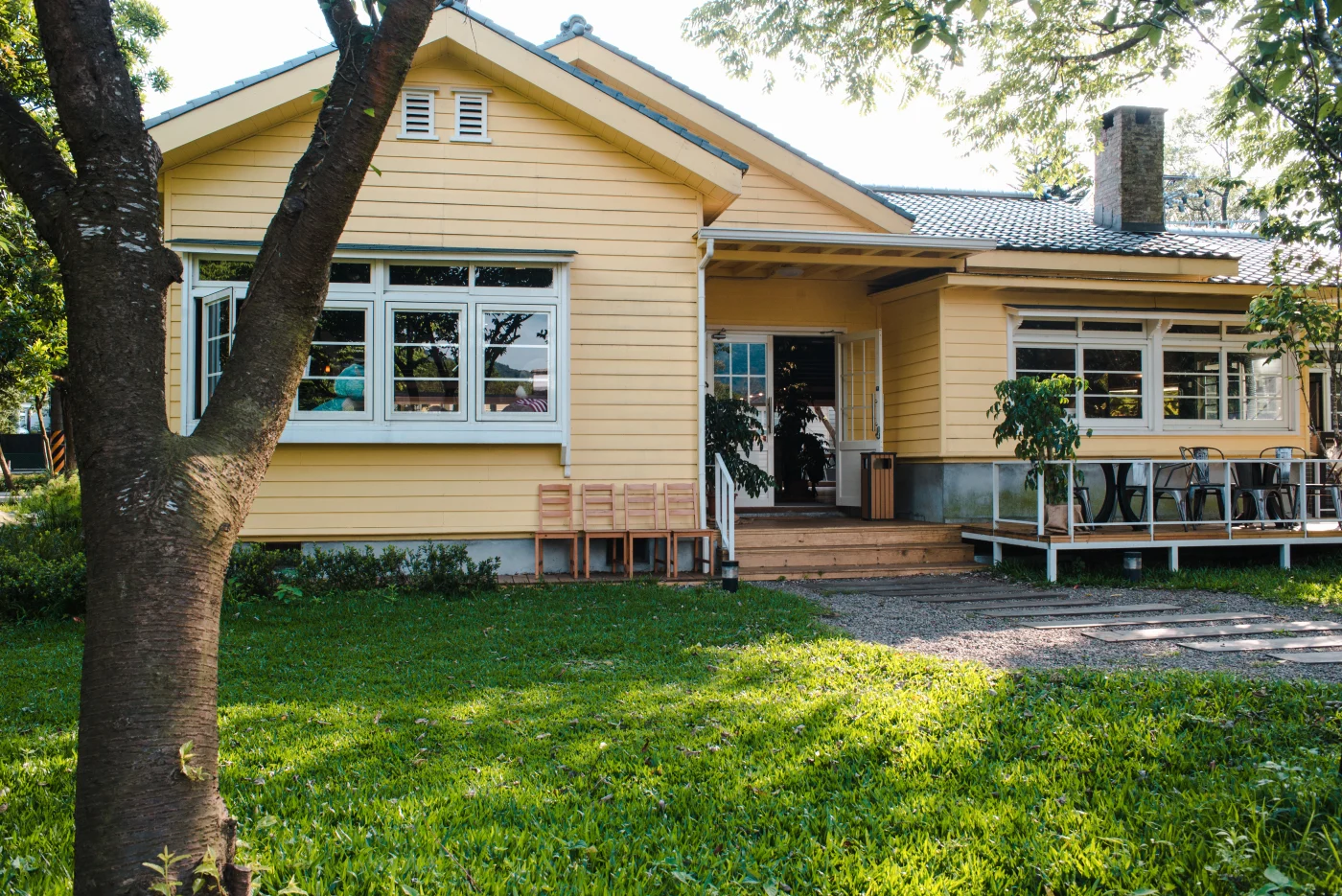This image features a house with yellow siding and white window trim. It is a small house with a beautiful grassy yard. There is a tall tree near the front of the yard.