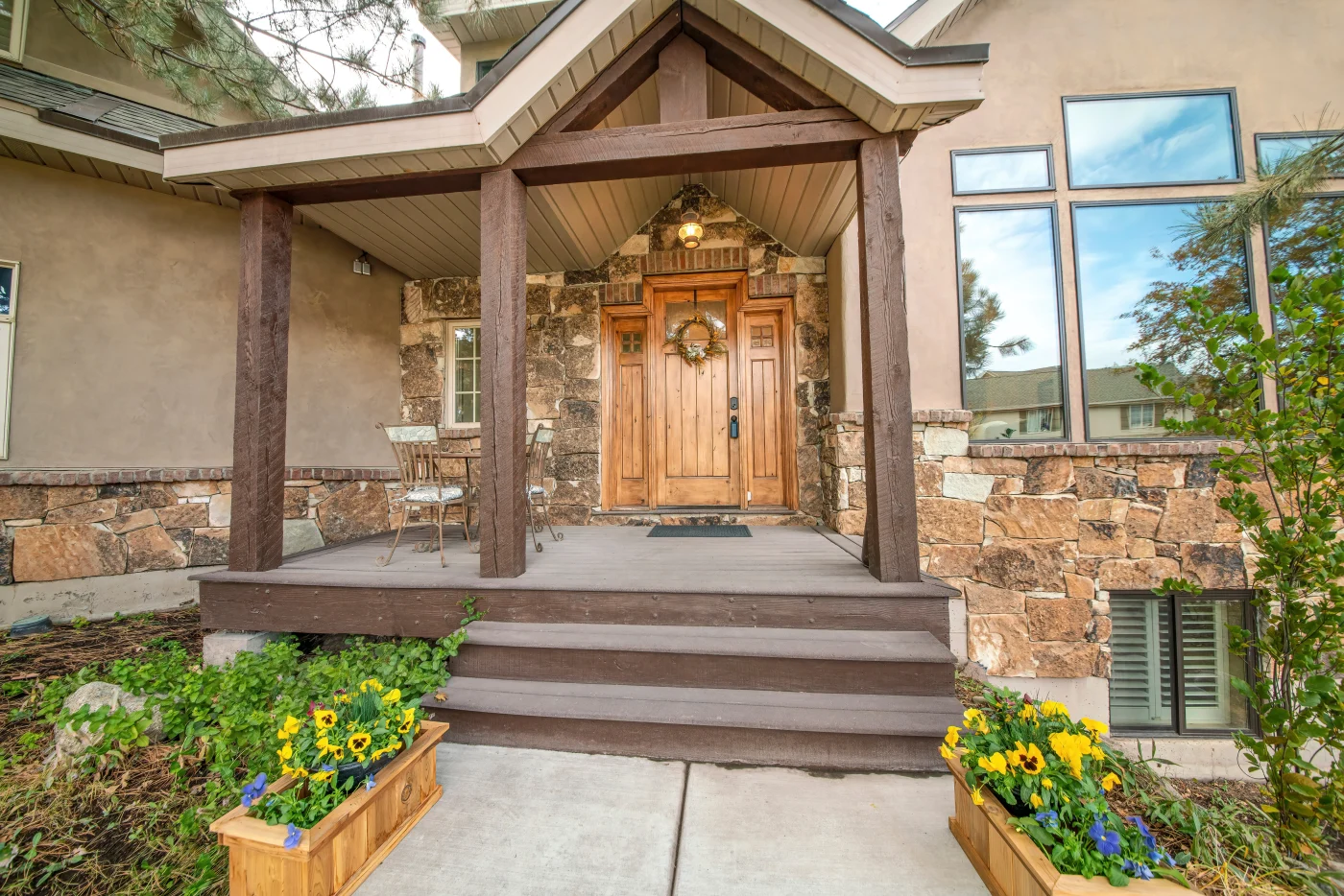 The entryway of a luxury home with a wood deck and two stairs, wood pillars and wood cover leading to a stylish single-entry wood door with wood sidelights and small little windows at the top, wrapped in engineered stone panel siding that goes about halfway up the home with stucco on the top half.
