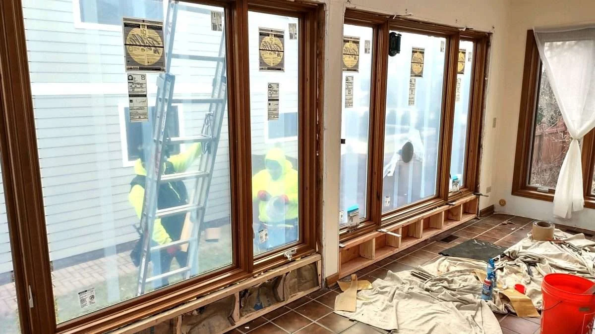 Close interior view of Peggy T.’s in-progress window replacement, focused on two side-by-side, multi-paned picture windows. The nearly completed short wall is visible below