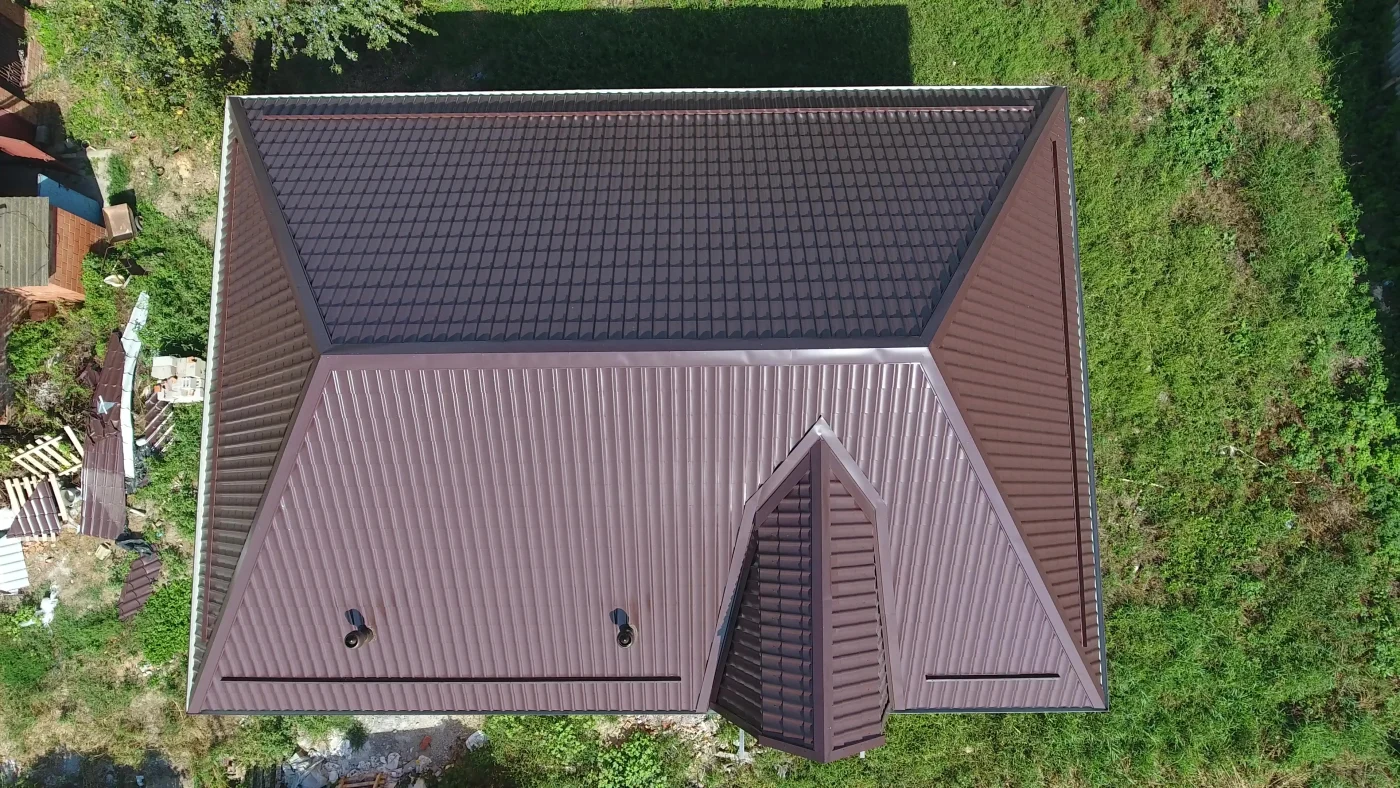 The image features a home in a grassy yard. The house has a brown metal roof with a wavy texture. With parts of metal roofing on the ground, it seems the roof was recently repaired.