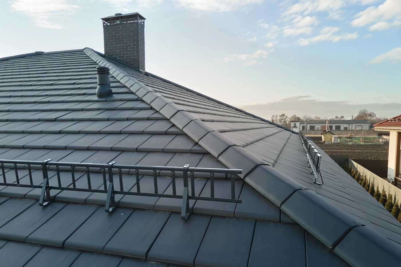 The image is of a roof with black slate and tile roof. It has a nice chimney. The home is in a rural area.