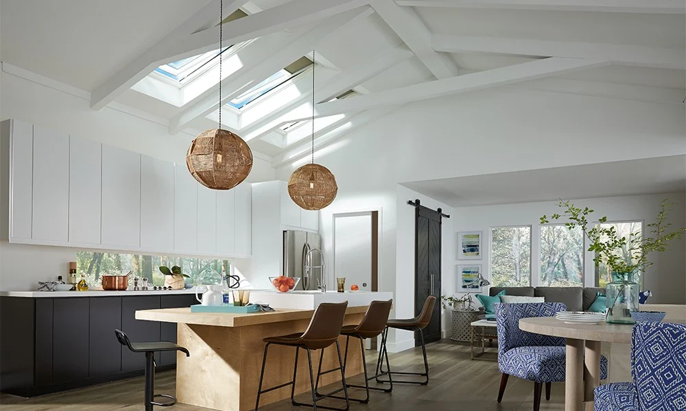 The inside of a modern, well furnished open-concept home, showing the kitchen with a wood island, white and red cabinetry, two earthy lighting fixtures, a couch to the far end below a set of three picture windows, a small dining table with colorful blue and white chairs, and three Velux skylights over the kitchen lighting the whole space.