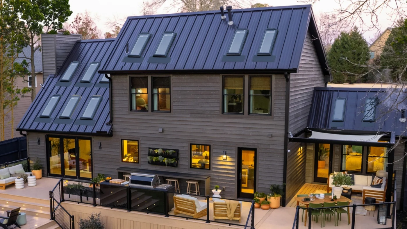 A large beautiful home with vertical plank siding and metal roofing, showing a total of 12 Velux skylights, and a variety of windows and three different entries. Also, a large deck with dark railing and deck furniture.