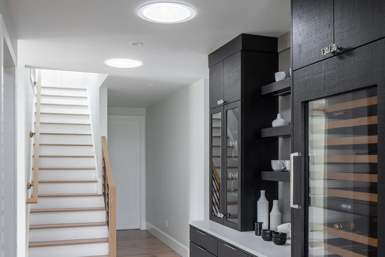 A look at an entryway and hallway to a home with a nice white staircase with wood accents going up, shiny wood flooring, two Velux sun tunnels lighting the area, and a large dark-colored hutch with different sections including one for wine.