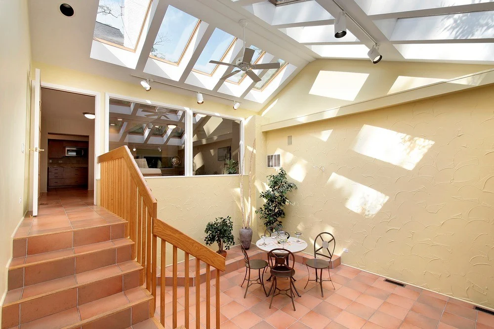 The inside of a large covered and enclosed front patio area with a small set of tiled stairs and wood railings, a small classic table with three chairs and settings, two large windows and an open door with white trim going into the house, and 10 Velux skylights covering the ceiling with lighting and a fan.