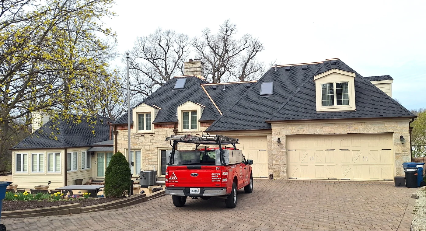 Apex Exteriors Offers An Experience-Driven Selection Of Exterior Brands & Products A luxurious home in the Chicagoland area with an Apex Exteriors truck in the driveway. The home is wide, with a dark shingle DaVinci roofing system and complex architecture. It has a combination of light brown or beige panel siding and trim, as well as different kinds of stone siding, many windows, two skylights, and two garages.