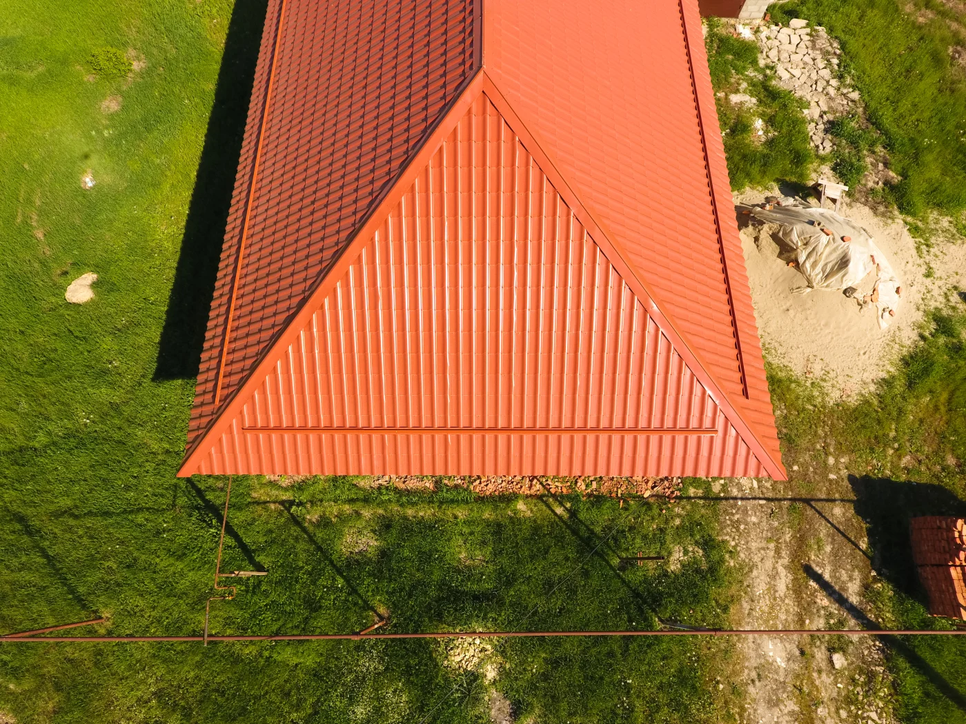 The image shows a grassy yard with a bit of dirt by the house. The house features a red metal roof with a raised line texture