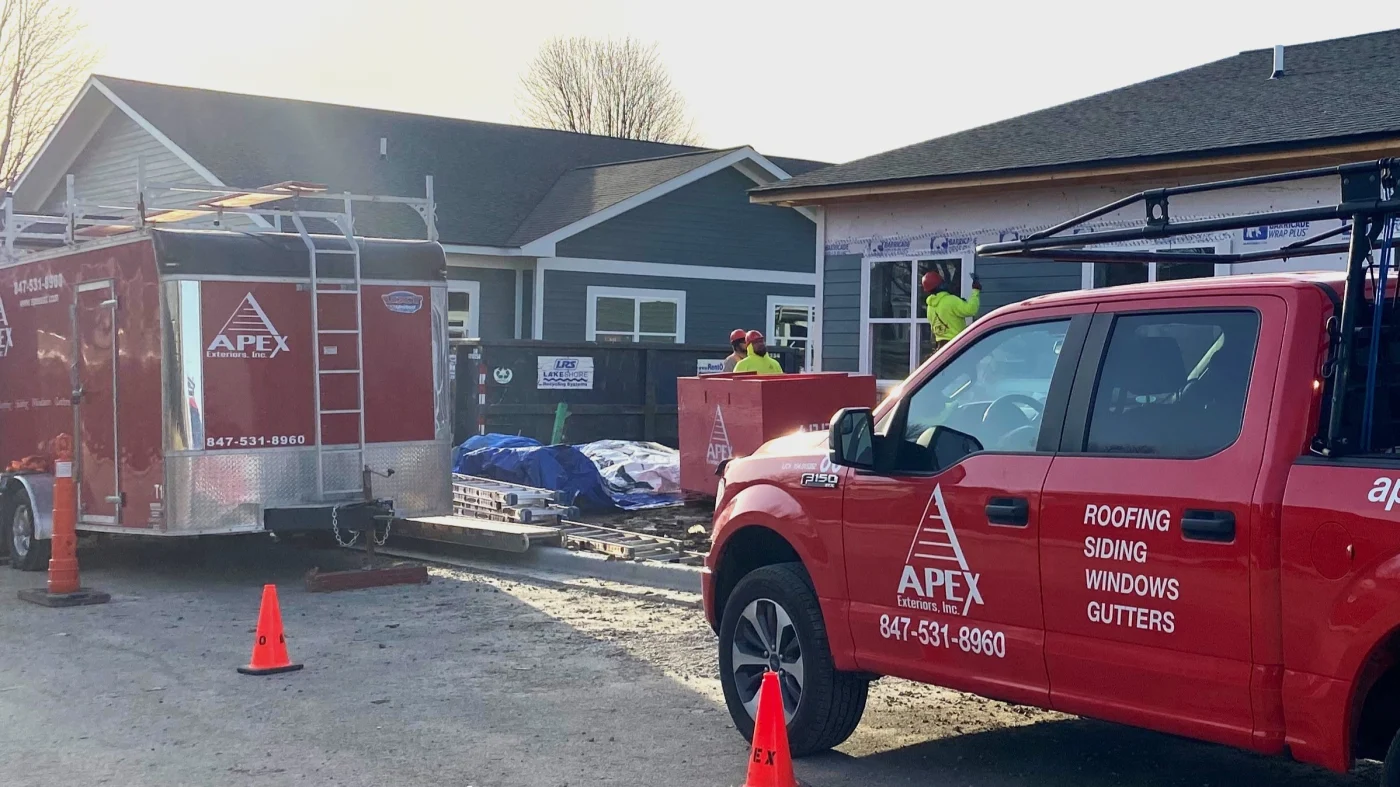 Two Apex Exteriors trucks are parked in front of houses as crews install new siding in Wheaton, IL by Apex Exteriors.