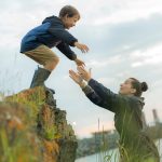 A mom holding out her arms to catch her son as he jumps off a small ledge in front of her.