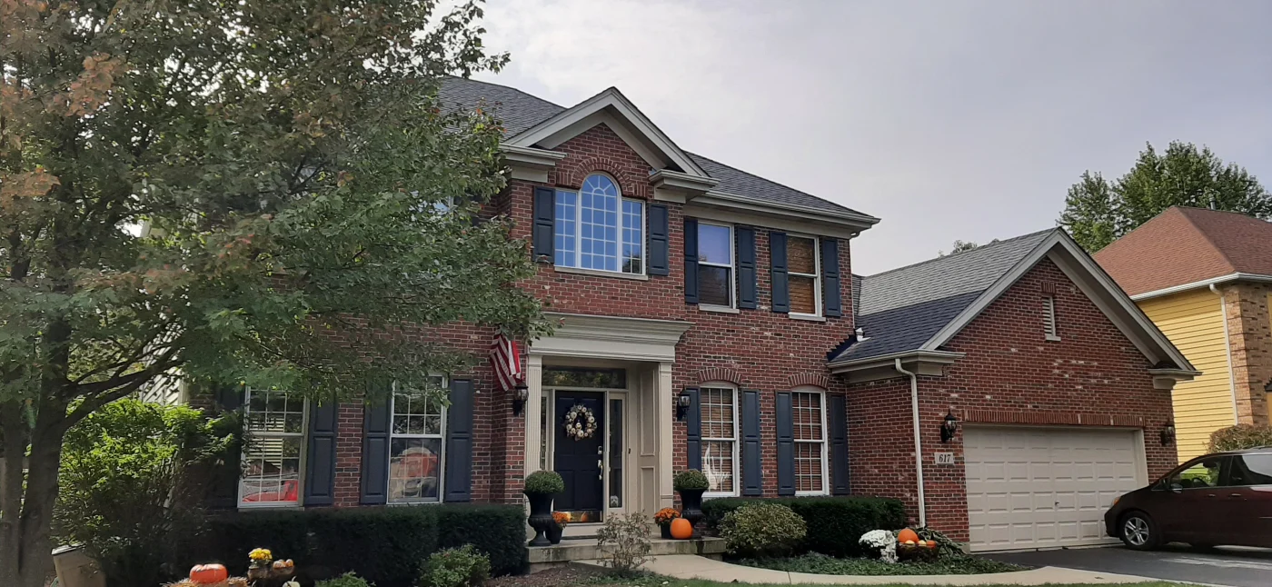 Brick two-story home with updated windows and entry door, dark shutters, and tidy landscaping, with fall pumpkins and front porch décor.