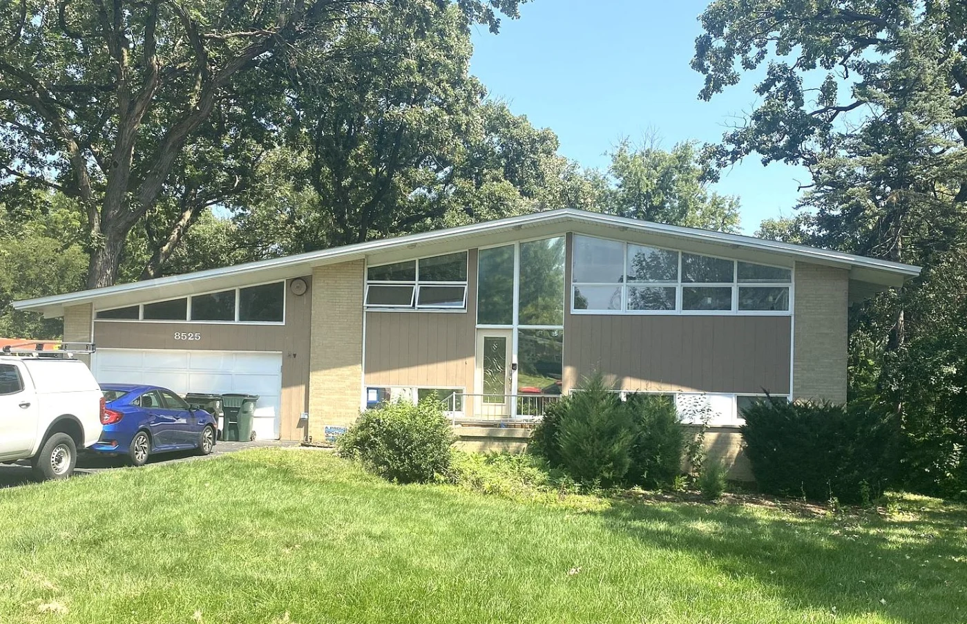 Mid-century modern home with expansive replacement windows, updated entry door, and an angled roof, surrounded by trees and a bright green front lawn.
