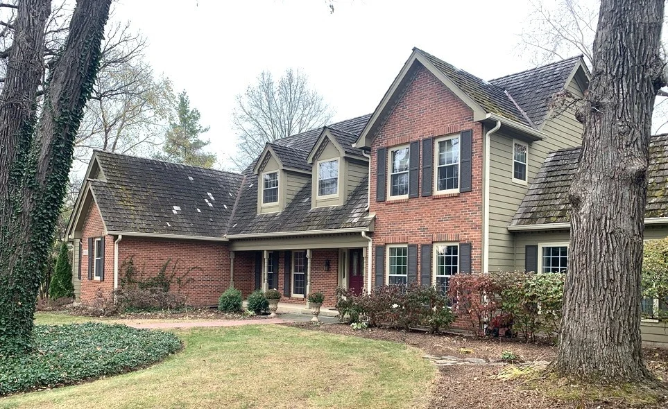 Two-story brick and siding home with classic double-hung windows, a red front door, weathered wood shake roof, and mature trees.