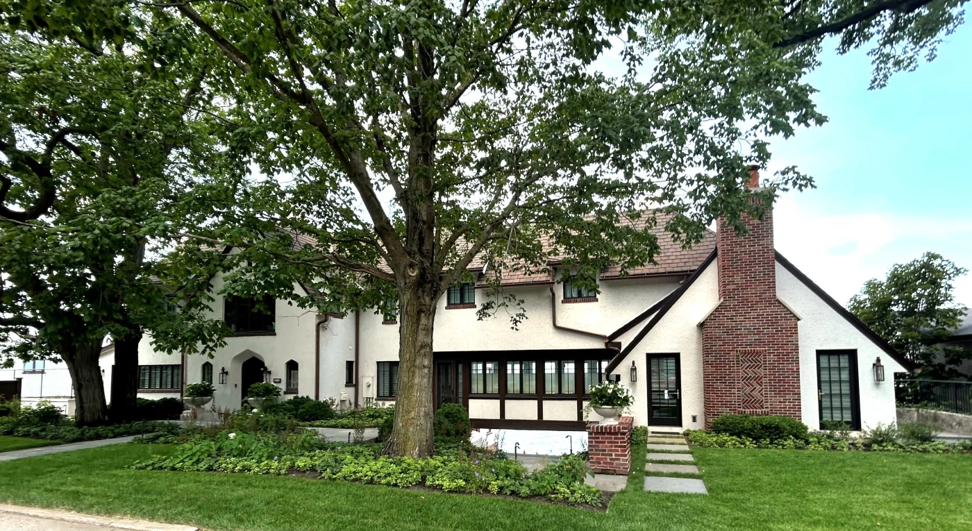Tudor-style home with white stucco exterior, brick chimney, black-framed windows, and landscaped front yard featuring mature trees and green lawn.