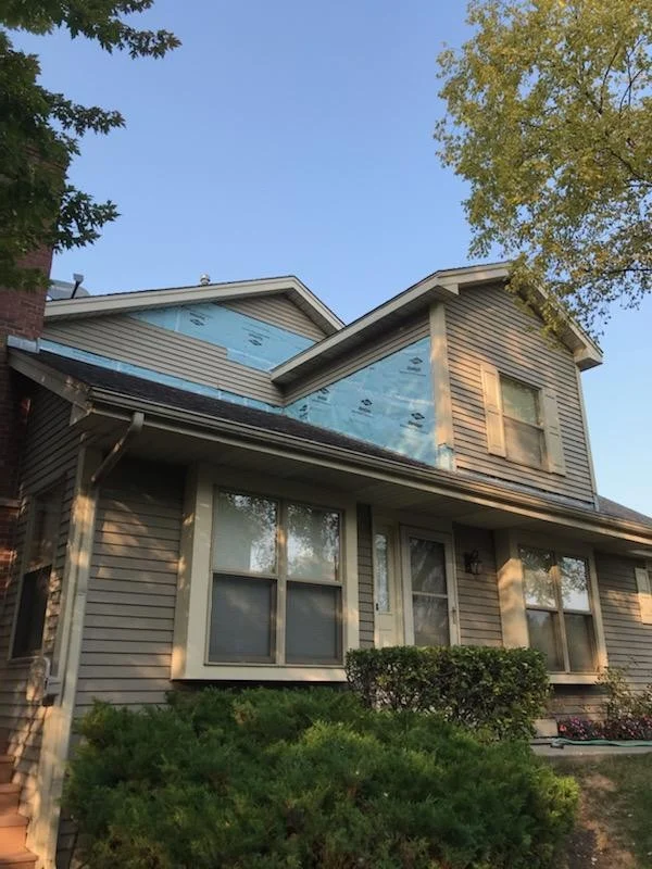 Two-story home with tan siding and white trim, showing partial exterior renovation with new windows installed and house wrap visible near the roofline.