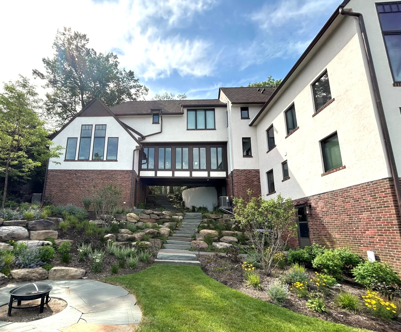 Rear view of a Tudor-style home with white stucco and brick exterior, large black-framed windows, elevated breezeway, stone steps, and landscaped backyard.