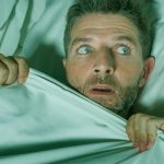A man in a bed, pulling up the covers in fear.