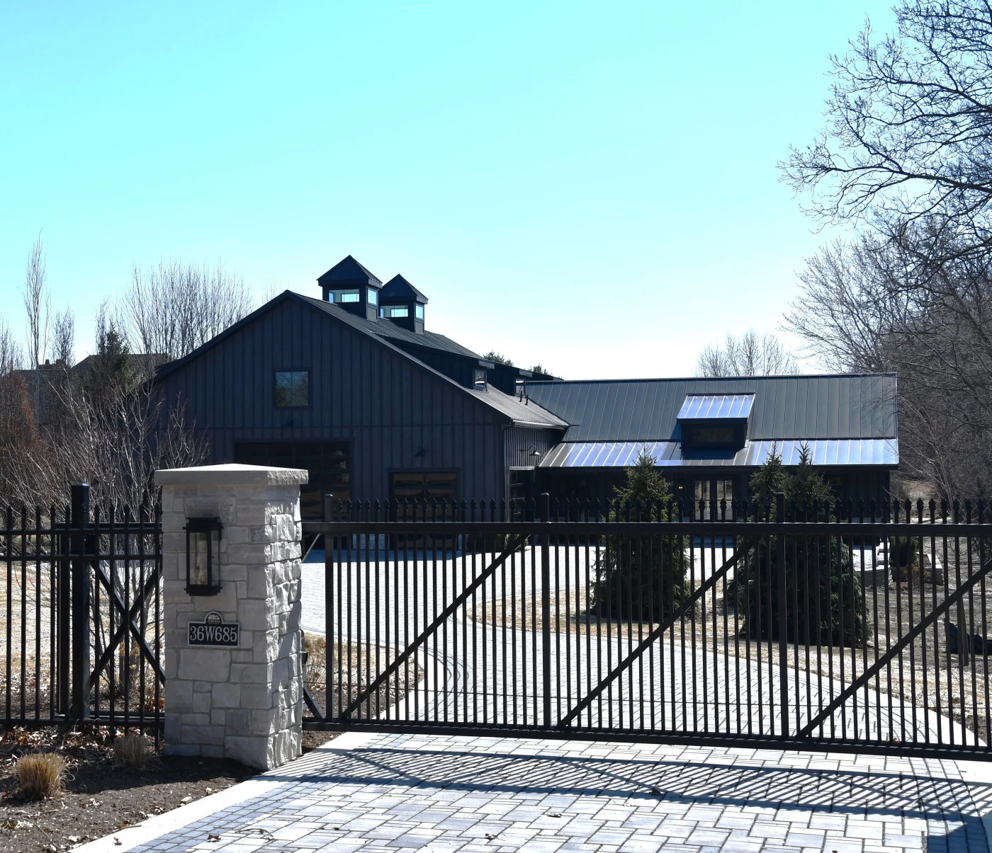 A modern black-clad home with multiple standing seam metal roofs, cupolas, and a gated stone entry, viewed from the driveway on a clear day.