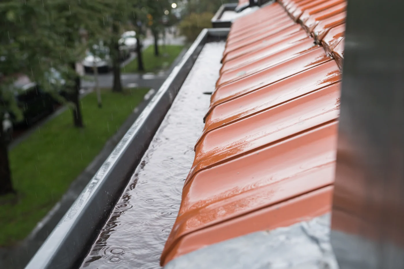 Awesome Gutter Replacement In Elgin, IL | Apex Exteriors A terra-cotta tiled roof on a rainy day. The visible gutters are partially filled with water. There are grassy yards, a sidewalk, trees, and a street visible in the background on the left side of the image.