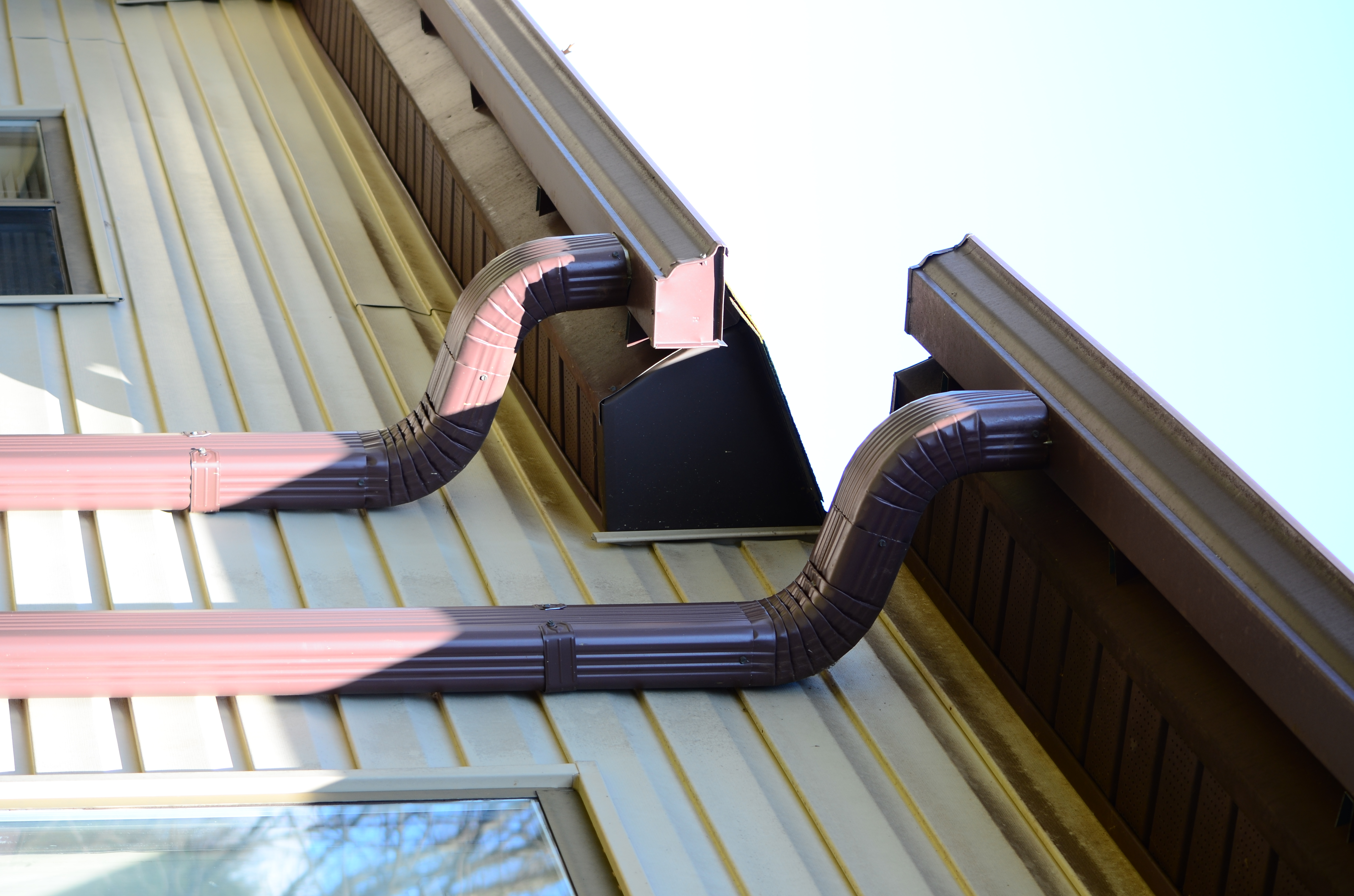The back outer wall of a home with cream siding, a brown roof, and gutters. The home is split-level, and two individual gutters and downspouts are pictured. There are portions of two windows visible, too.