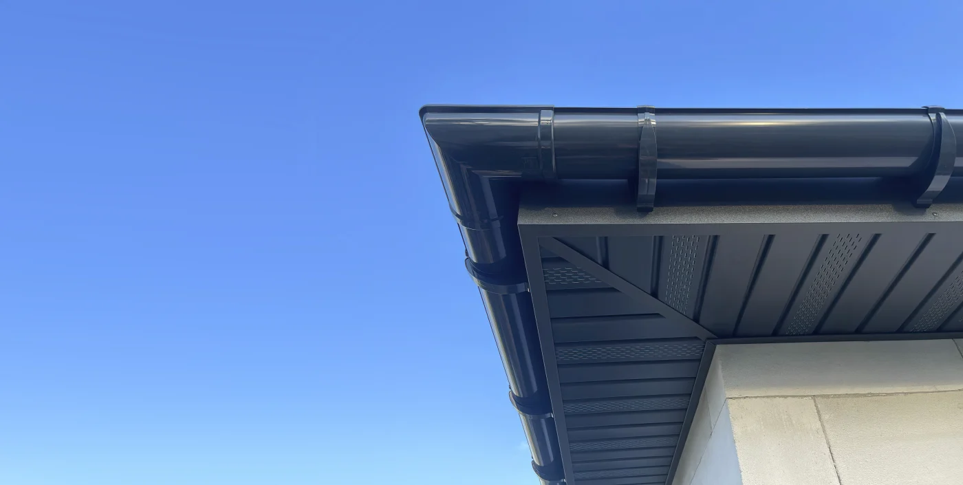 Broad Soffit And Fascia Replacement In Elgin, IL | Apex Exteriors A corner of a home's roof and gutter system. There is no downspout visible, but the gutters, soffit, and fascia are dark colored, while the house is white. The blue sky is bright in the background.