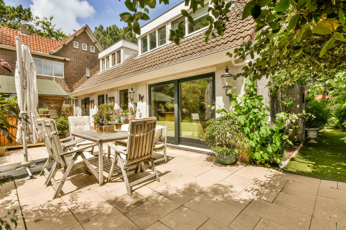 A home’s backyard patio, looking toward the house. There is a wooden table and four wooden chairs, and a closed umbrella on the patio. The patio tiles are tan colored, and so are the home’s shingles. The door leading out to the patio is large and made of glass panes, and has a black frame. There is a lot of greenery surrounding the house, including bushes, trees, and grass.
