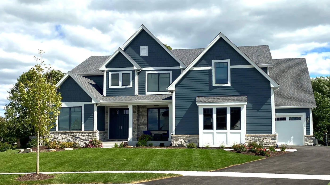 Dependable Gutter Installation | Wayne, IL | Apex Exteriors Inc. Two-story home with dark blue siding and white trim, featuring a covered entry, stone accents, and a visible gutter system along the roofline, with a neatly landscaped front yard beneath a cloudy sky.