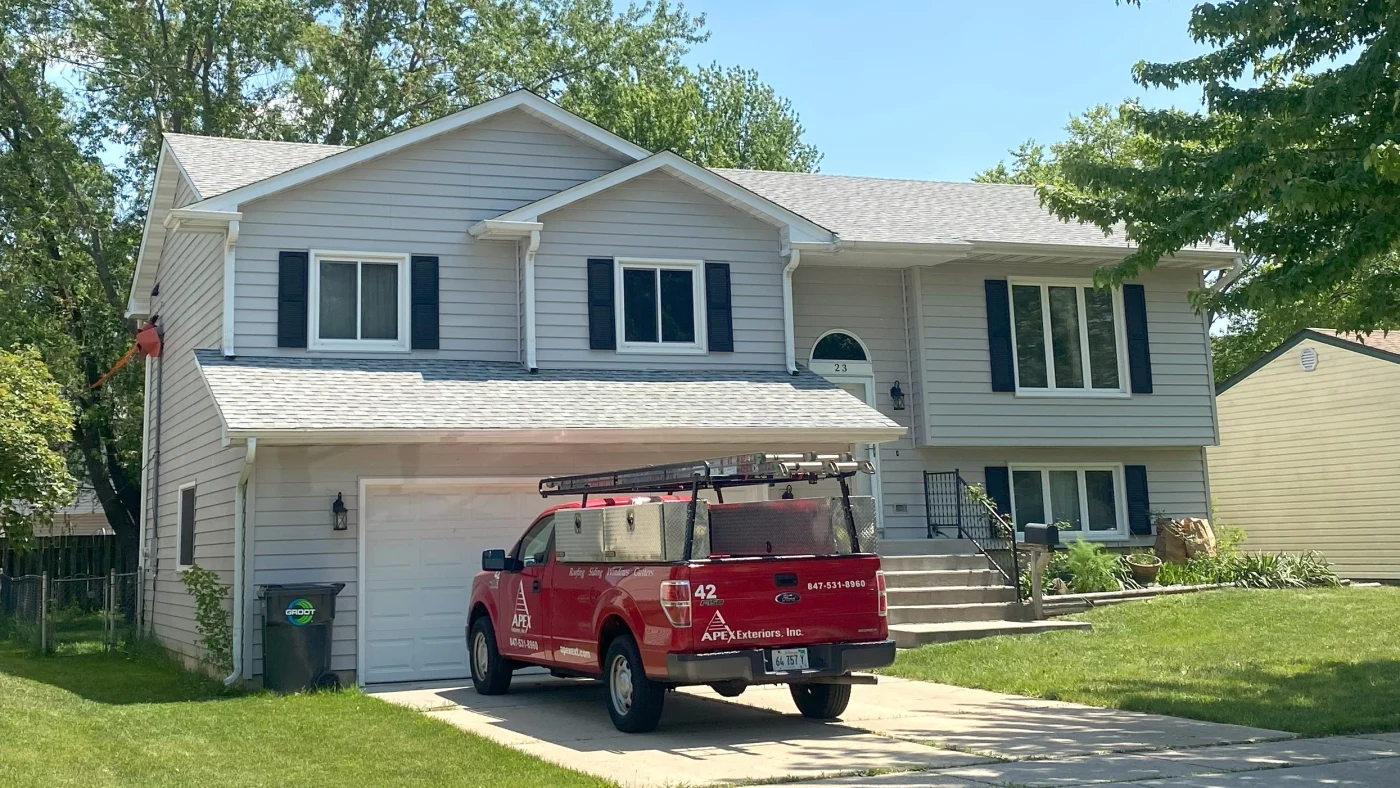Red Apex Exteriors pickup truck parked in the driveway of a two-story home with light siding and a newly updated asphalt shingle roof, highlighting clean roof lines and proper installation.