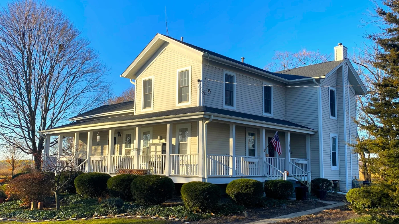 Dependable Window Installation | Wayne, IL | Apex Exteriors Inc. Two-story home with light vinyl siding, multiple double-hung windows, and a dark shingle roof, featuring a full wraparound porch with white railing and clean landscaping under a clear blue sky.