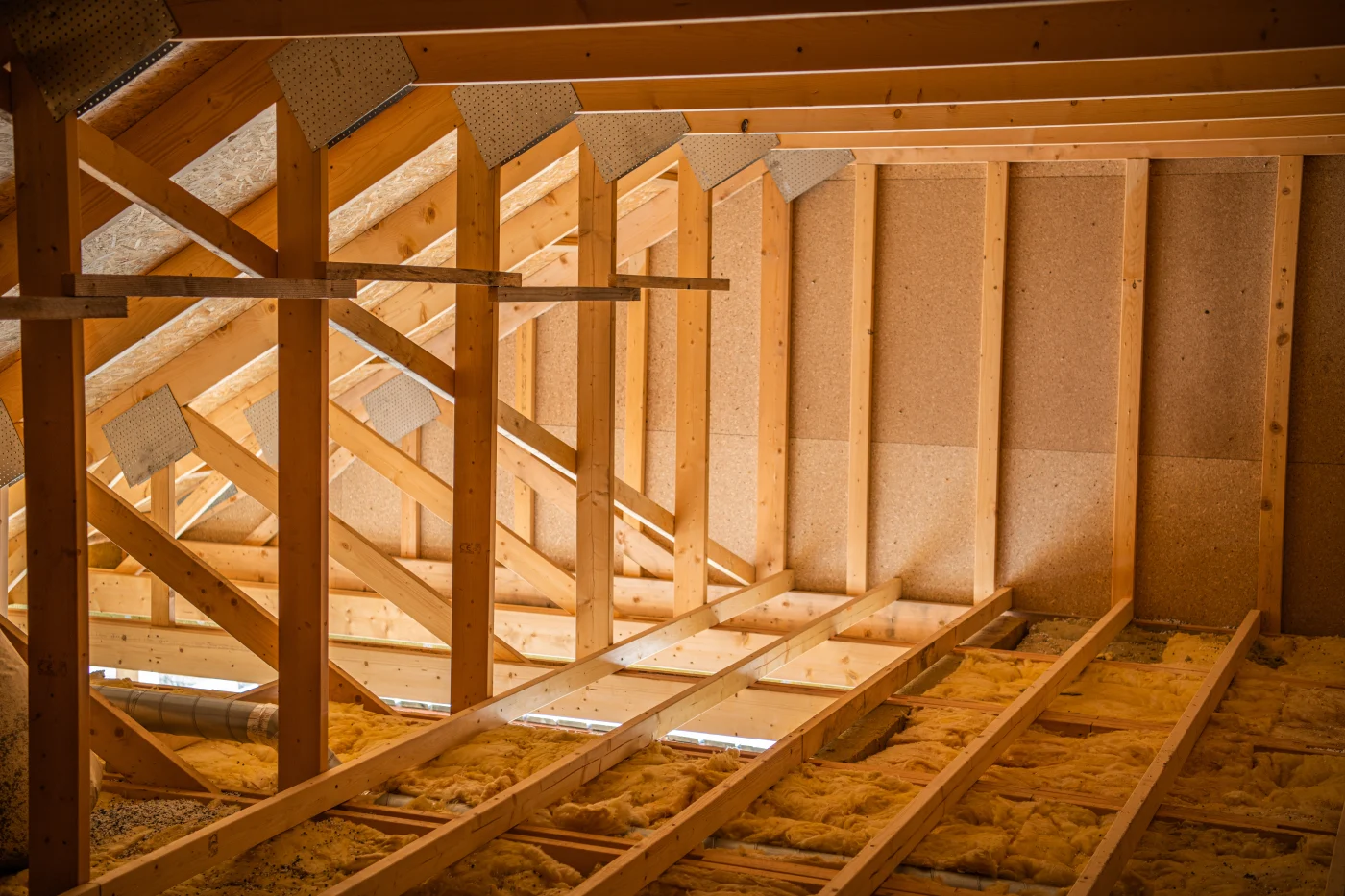 An unfinished attic with exposed wooden support, floor, and ceiling beams. There is insulation between some of the floor beams. Sunlight illuminates the attic from below, as it comes in through the open portion of the floor.