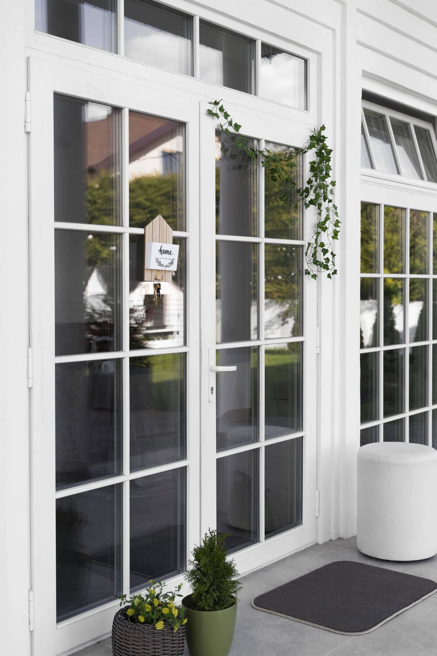 A close-up view of a set of French doors with white trim and framing. Each of the two doors is made of 8 square panes of glass. There is a small sign that says 'home' in black script hanging on the left door, and an ivy vine draped against the right.
