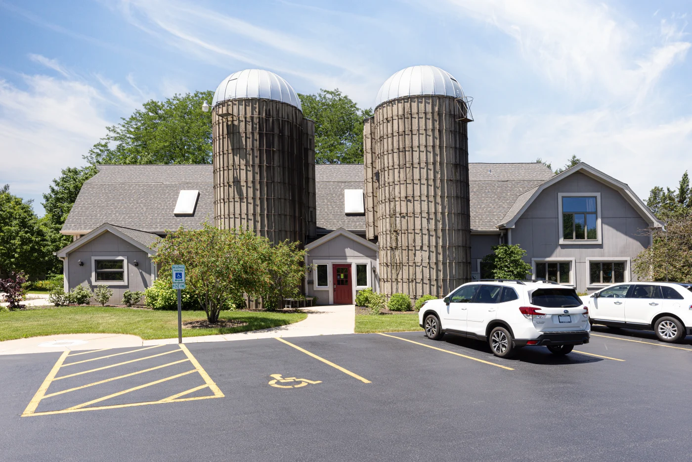 A front view of a gray commercial building with two tall concrete silos, a red entry door, paved parking lot with white SUVs.