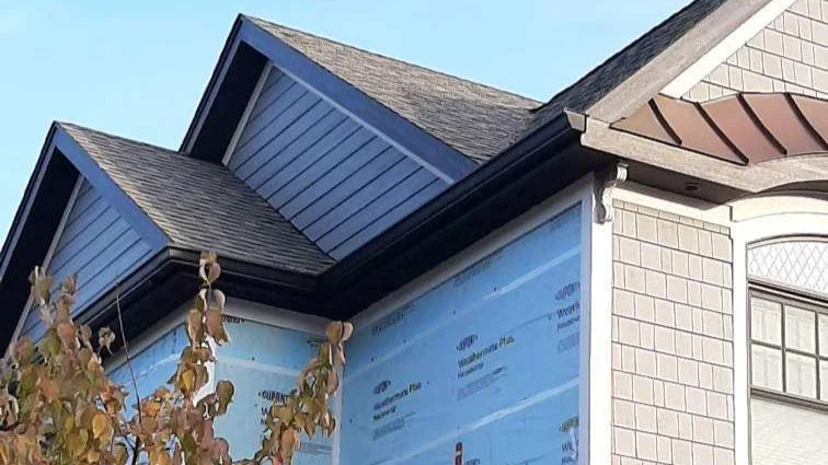 Expert Gutter Installation | Wayne, IL | Apex Exteriors Inc. Close-up of a home exterior under construction, showing newly installed roofline with black gutters and downspouts, light siding details, and protective wrap visible beneath the eaves.