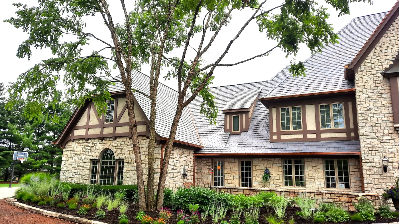 Expert Roofing Contractor | Wayne, IL | Apex Exteriors Inc. Elegant stone home with a steep-pitched composite or slate-style roof, featuring intricate rooflines, dormers, and detailed trim set against mature landscaping.