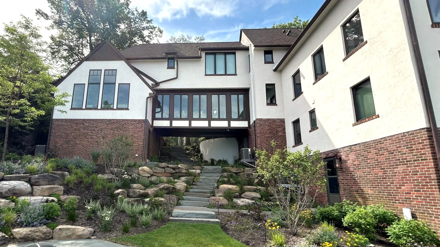 Expert Window Installation | Wayne, IL | Apex Exteriors Inc. Multi-level home with stucco and brick exterior, featuring large grid-style windows, a window-lined enclosed bridge between sections, and landscaped stone steps leading up through a terraced yard.