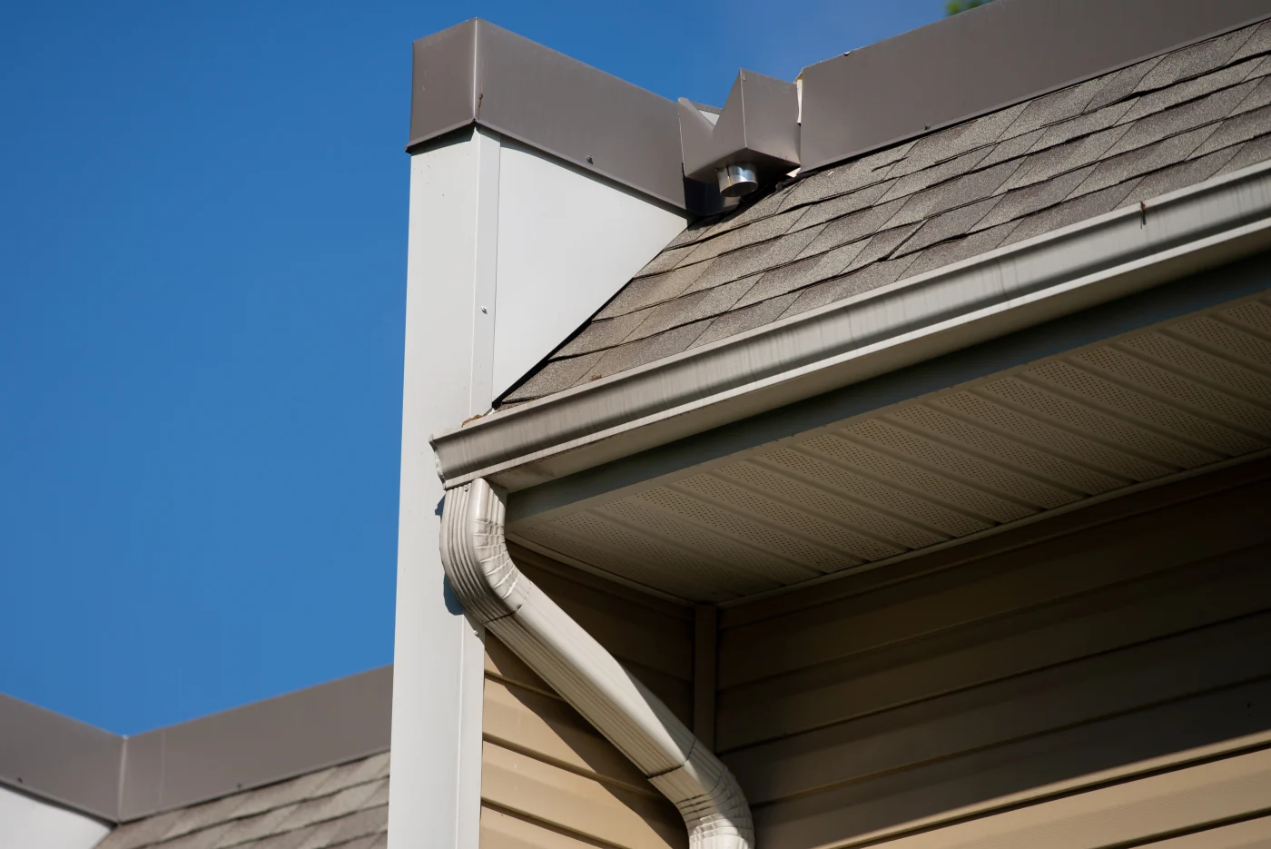 Part of a home's roof, featuring brown and tan shingles, light brown siding, and a white gutter system. The bright blue, cloudless sky is visible behind the house.