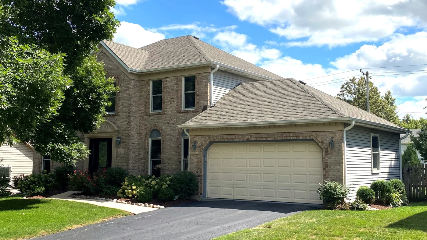 Outstanding Roofing Contractor | Elgin, IL | Apex Exteriors Inc. Two-story home with tan shingle roof, brick exterior, gray siding, and attached garage on a suburban lot.