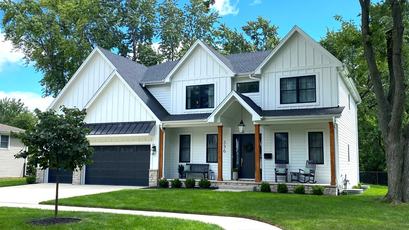 Outstanding Window Installation | Elgin, IL | Apex Exteriors Inc. Modern two-story home with white vertical siding, black-framed windows, and a dark shingle roof, featuring a covered front porch with wood columns and a two-car garage.