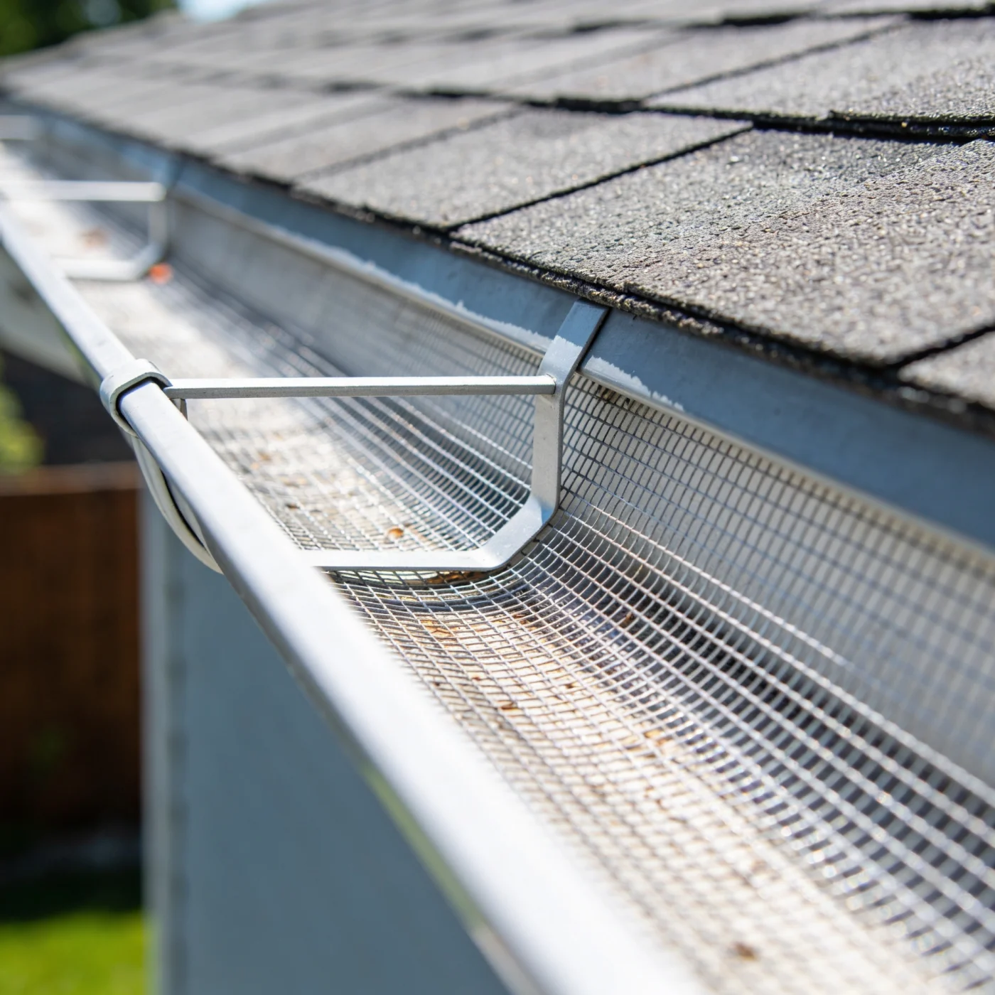 Practical Gutter Guards In Elgin, IL | Apex Exteriors A view of metal gutters with a screen installed inside. The roof is gray shingles, and the siding on the home is a light blue. You can see part of a tall wooden fence and a green grass lawn on the left of the image.