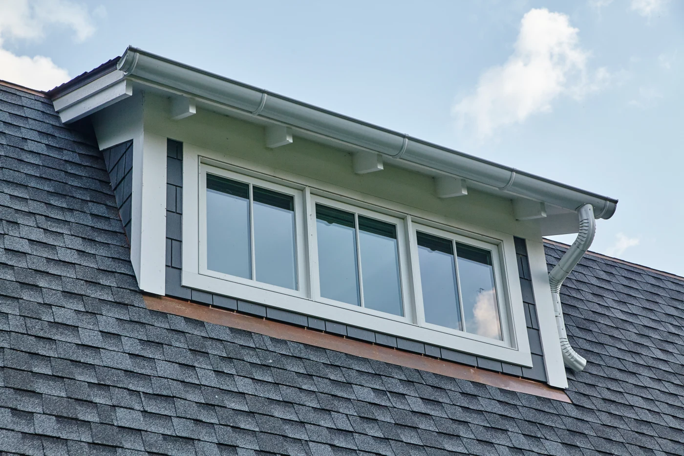 A home's dormer window with three individual windows arranged side by side. The gutters and trim are white, and the roof is a mix of light and dark gray shingles.
