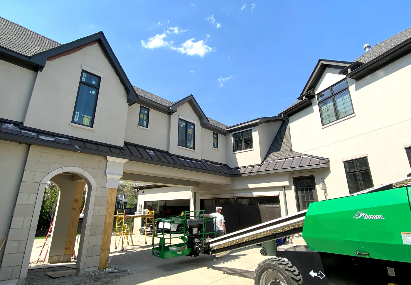 A modern black-clad home with multiple standing seam metal roofs, cupolas, and a gated stone entry, viewed from the driveway on a clear day.