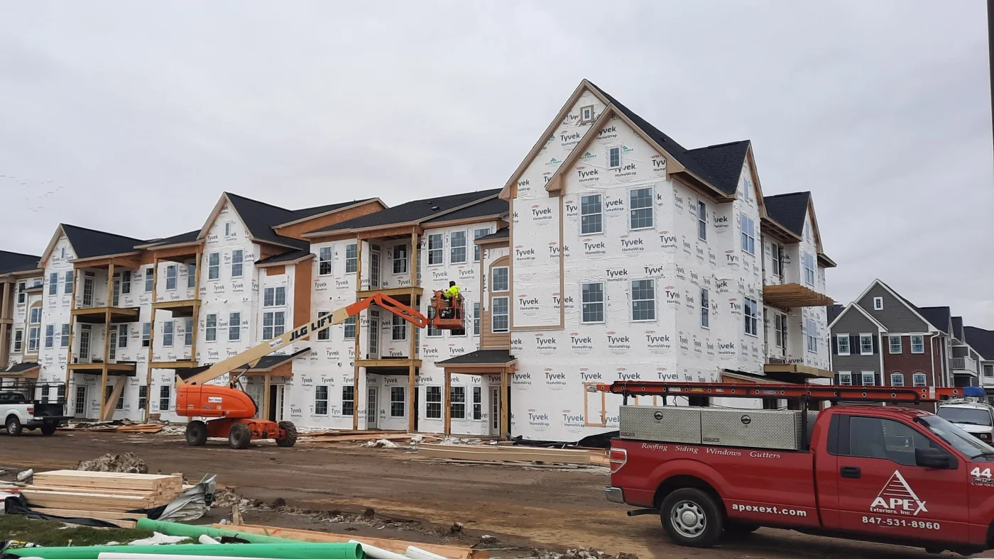 Remarkable Commercial New Construction | Elgin, IL | Apex Exteriors Inc. Large multi-family building under construction with Tyvek-wrapped exterior, installed windows, and framing details, featuring Apex Exteriors truck and lift equipment at the site.