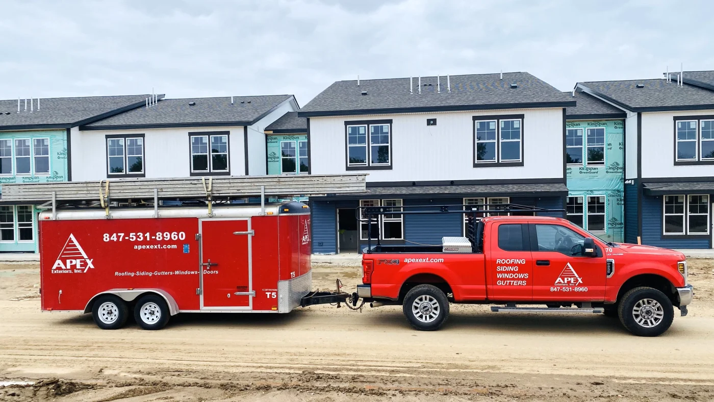 Remarkable Exterior Remodeling Services | Elgin, IL | Apex Exteriors Inc. Red Apex Exteriors truck and trailer parked in front of a multi-family housing development under construction, with newly installed siding, windows, and roofing materials visible across the building exteriors.