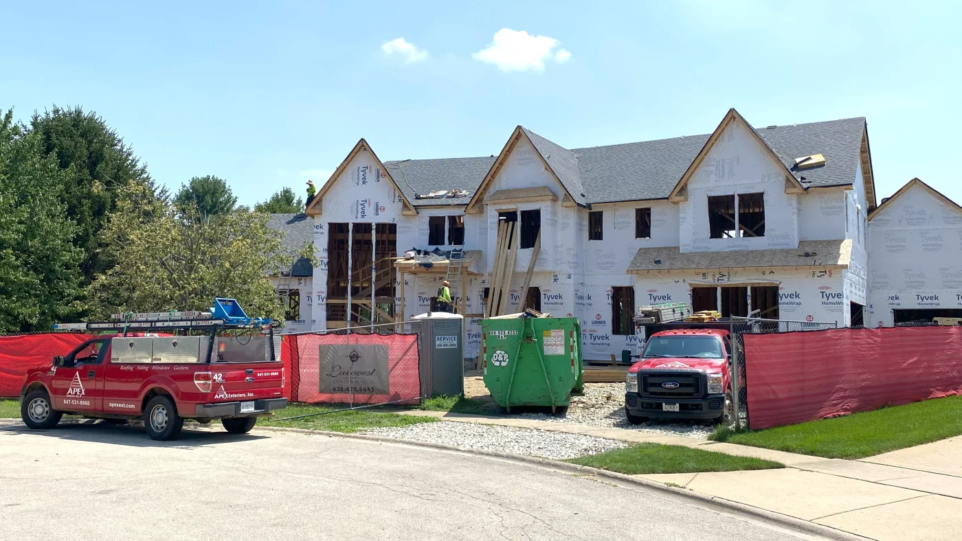 Remarkable New Home Construction | Elgin, IL | Apex Exteriors Inc. New residential construction in progress with Tyvek-wrapped exterior, dark shingle roof, and multiple gables, featuring Apex Exteriors trucks parked at the jobsite.